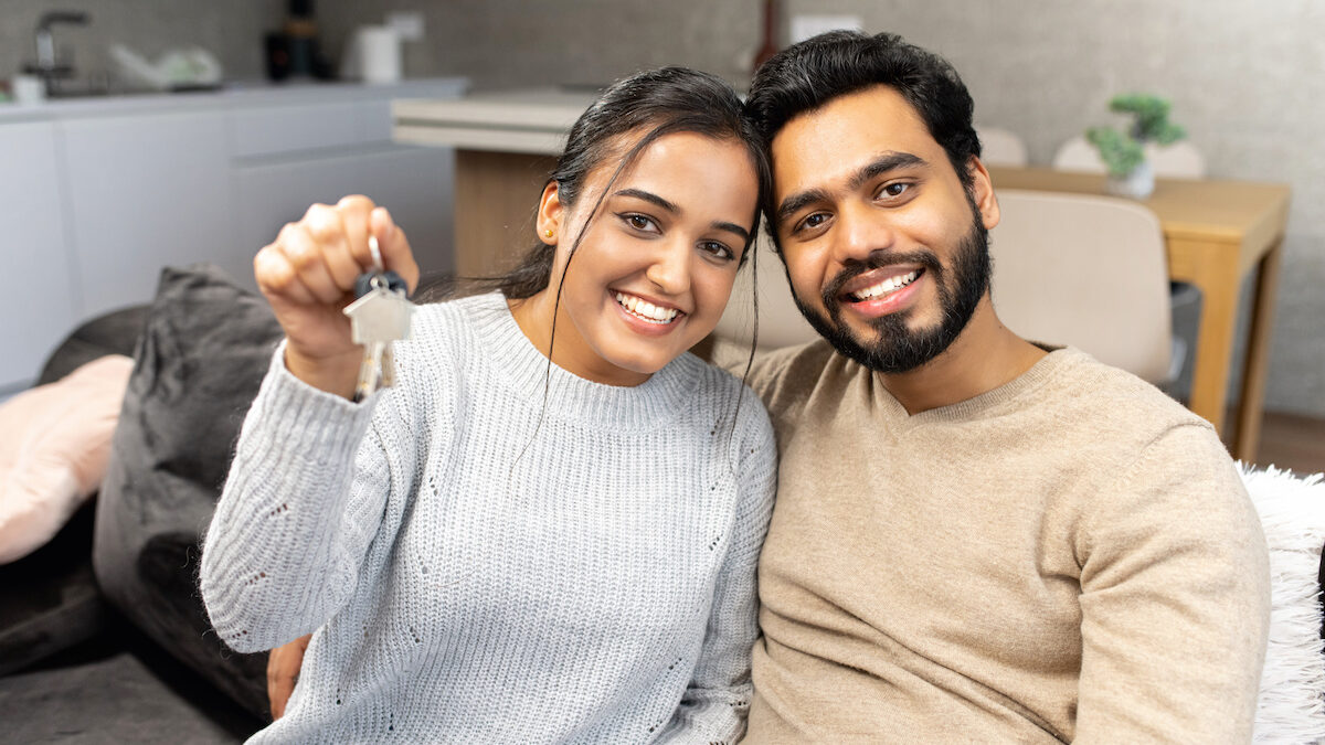 Young married couple smiling cheerfully and showing keys from a new apartment, hugging and looking at camera, standing in the modern kitchen of a new home